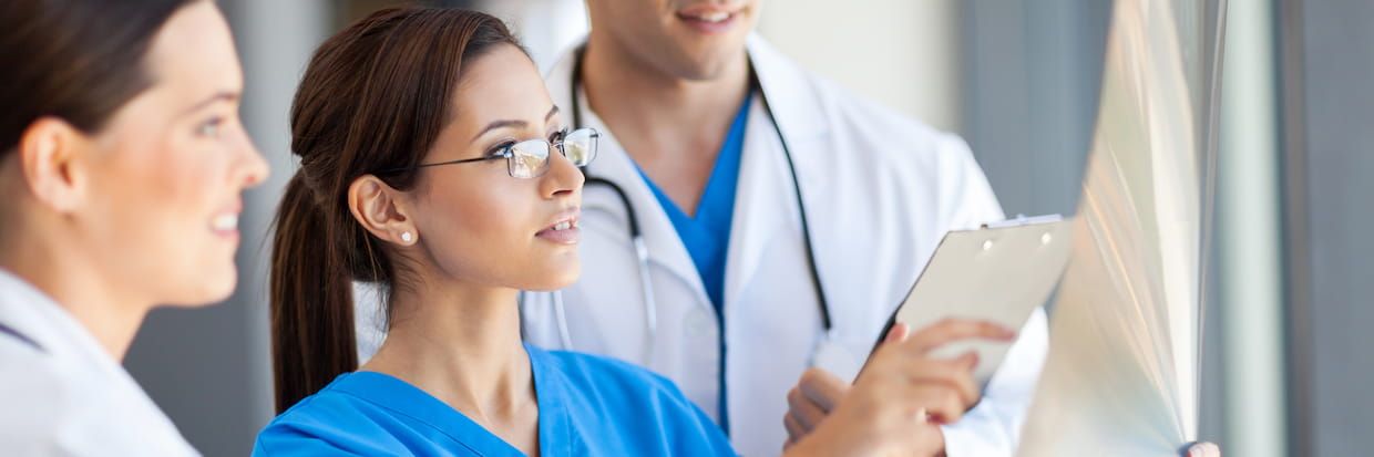 Three healthcare professionals, two women and one man, discussing medical reports, one holding a clipboard and another looking at an x-ray. They appear engaged and focused on health education in a bright clinical setting.