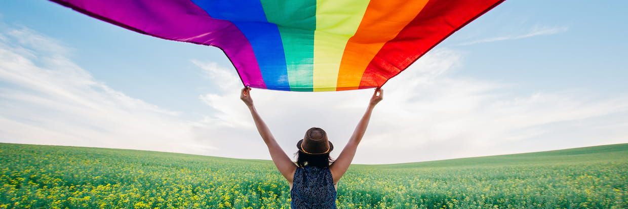A person in a straw hat and sleeveless top holds a large rainbow flag, symbolizing LGBTQIA community pride, above their head in a vibrant yellow flower field under a clear blue sky.