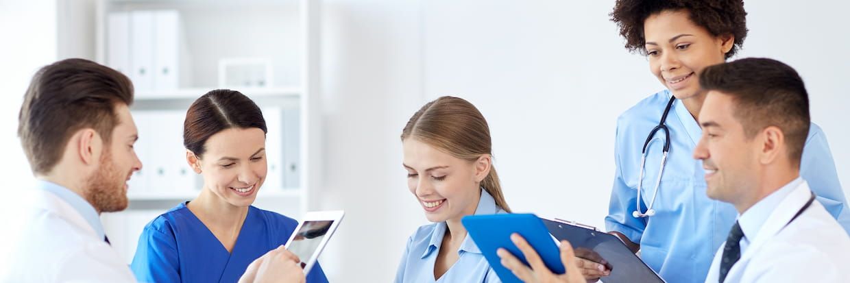 A group of five healthcare professionals, including two men and three women with diverse ethnicities, discuss medical information using a tablet and documents in a bright office, focusing on student loan forgiveness for nurses.