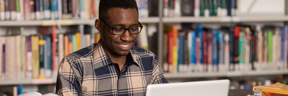 A smiling man in a plaid shirt using a laptop in a library with shelves of colorful books in the background.