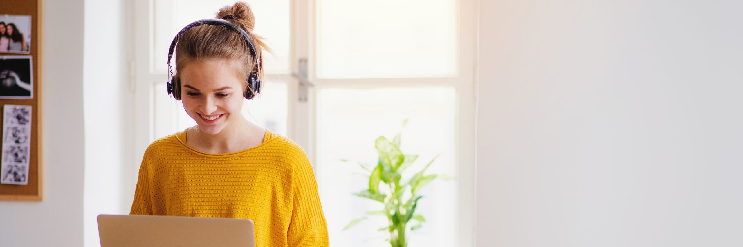 A young woman wearing headphones uses a laptop in a brightly lit room with pictures on the wall and a plant nearby, smiling as she looks at the screen.