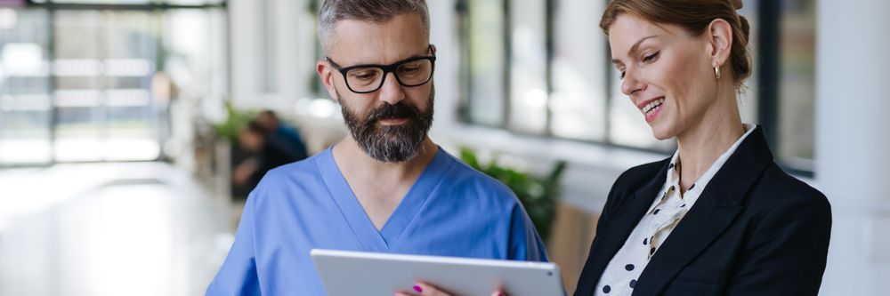 A male nurse in blue scrubs and a female doctor in a suit review information on a tablet together in a hospital corridor.