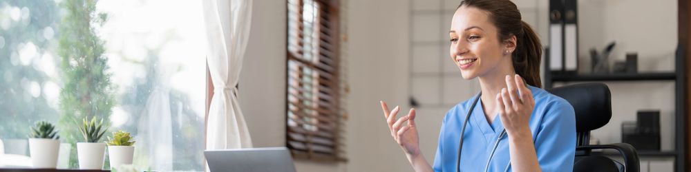 A smiling healthcare professional wearing a blue uniform and stethoscope engages in a video call on a laptop. She is seated in a bright room with potted plants on the windowsill and shelves in the background, discussing CNA renewal requirements for every state.